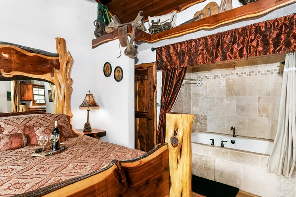 Un dormitorio con una cama de madera, una mesita de noche con lámpara, un espejo grande y un baño con bañera y paredes con azulejos.