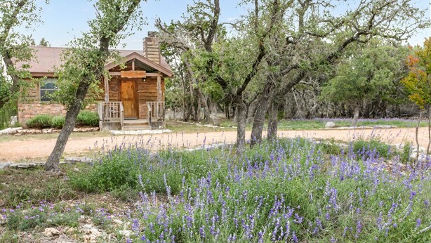 Una cabaña rústica con porche de madera y chimenea de piedra, rodeada de un jardín con flores moradas y árboles.