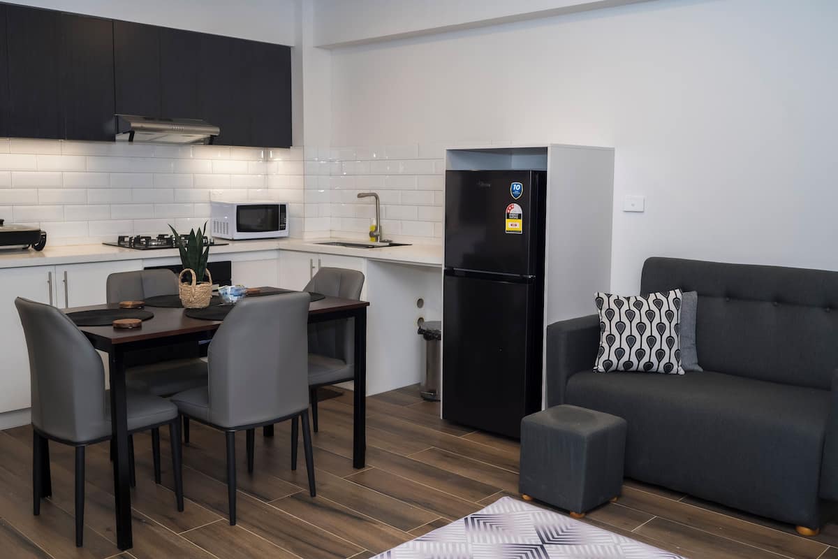 A modern kitchen with dark cabinets, a microwave, a sink, and a refrigerator. There is a dining table with chairs and a grey sofa with a patterned pillow.