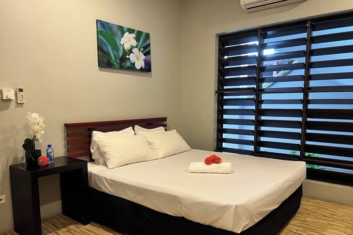A neatly made bed with white linens, a wooden headboard, and a single red flower on the pillow. A small table with a vase of white flowers and a water bottle is beside the bed.
