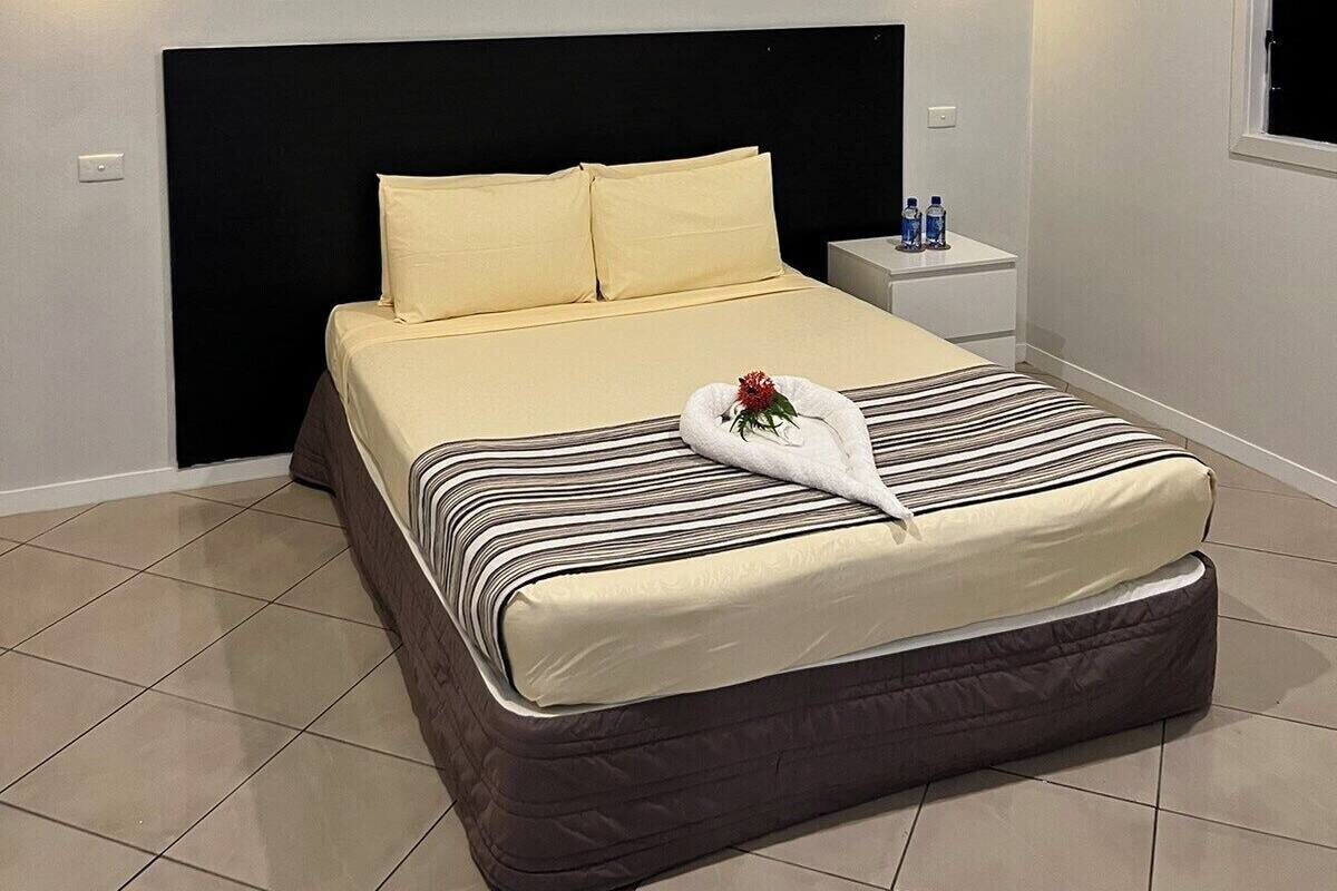 A neatly made bed with a black headboard, two bedside tables with water bottles, a ceiling fan, and an air conditioning unit.