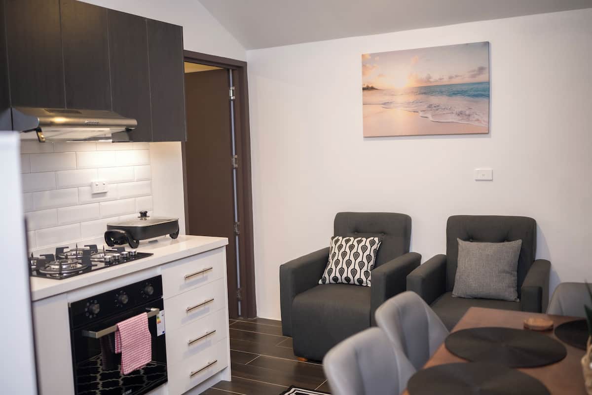 A modern kitchen with white cabinets, a stove, and a built-in oven. There is a dining area with a table and chairs, and a gray armchair with a patterned pillow. A painting of a beach scene hangs on the wall.