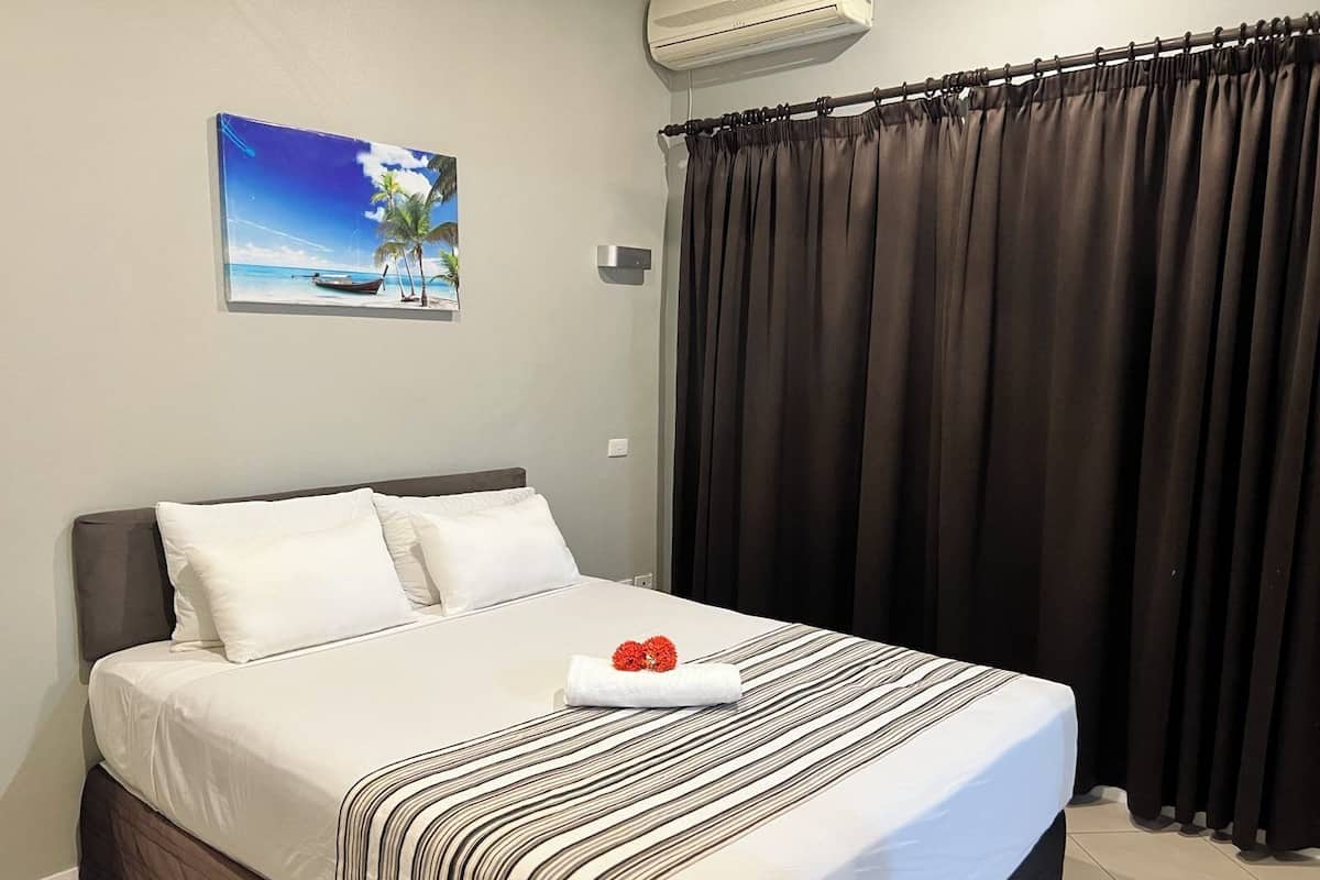 A neatly made bed with white linens and a striped blanket, a picture of a beach scene on the wall, and a dark curtain covering a window.