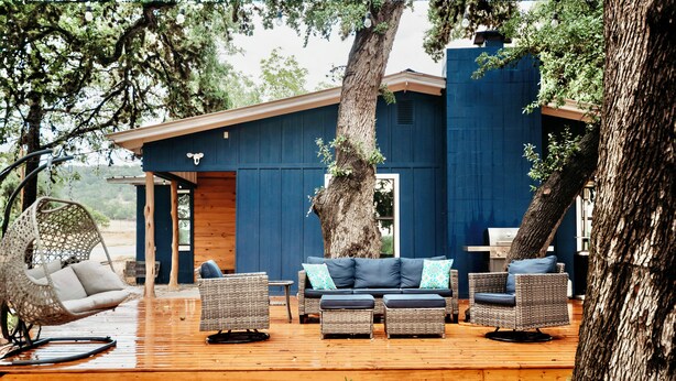 Una terraza de madera con muebles de mimbre, una silla colgante y un edificio azul con chimenea.