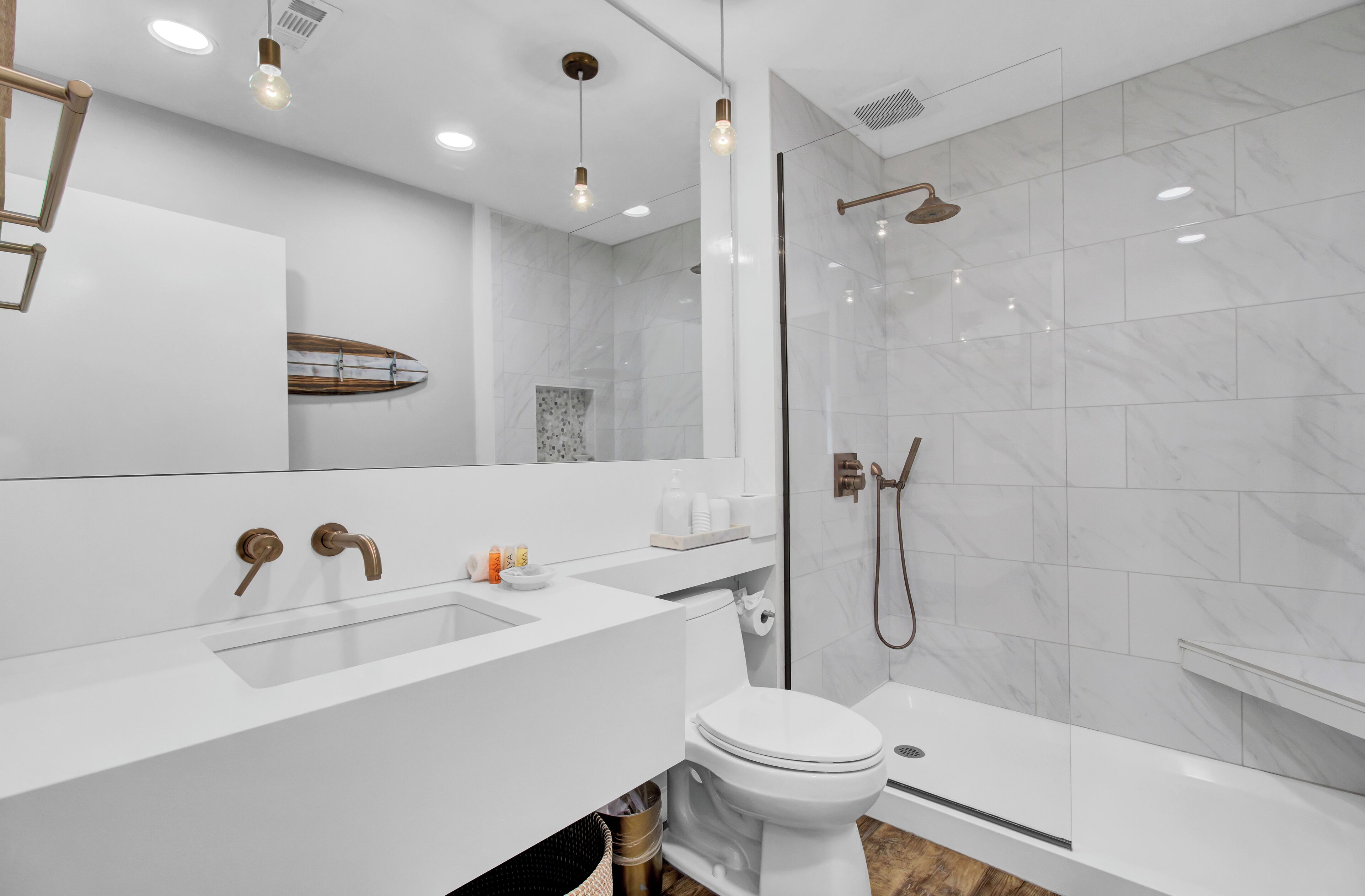A modern bathroom with a white sink, toilet, and shower. It features a large mirror, recessed lighting, and a surfboard decoration on the wall.