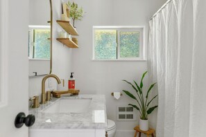 A bathroom with a marble sink, gold faucet, wooden shelves, a potted plant, and a window with frosted glass.