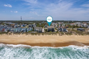Vista aérea de una zona residencial costera con casas a lo largo de la playa, cielo despejado y un tanque de agua al fondo.