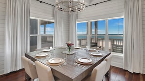 Comedor con una gran mesa de madera dispuesta para seis personas, cortinas blancas y vistas al océano.