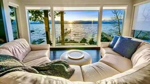A modern living room with a large window overlooking a lake, a beige sofa, and a small round table.