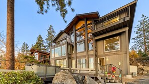 A modern multi-story house with large windows and a balcony, surrounded by trees and rocks.