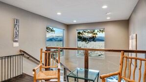 A modern interior with a wooden staircase, two wicker chairs, a glass table, and large windows offering a view of a lake and mountains.