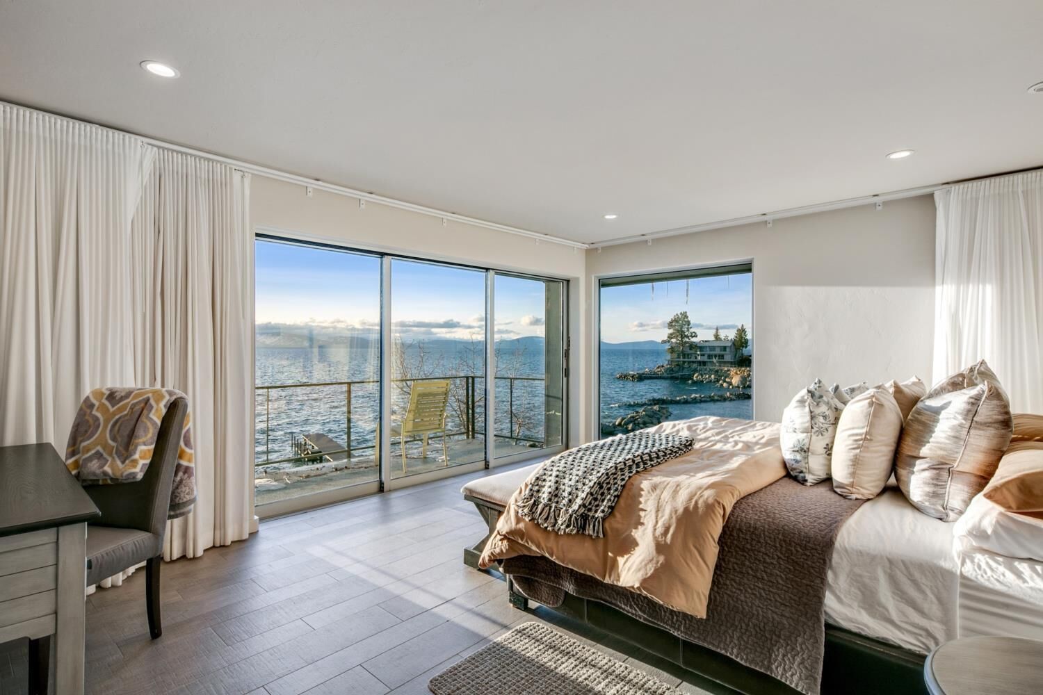 A modern bedroom with a large bed, a desk, and a view of the sea through floor-to-ceiling windows.