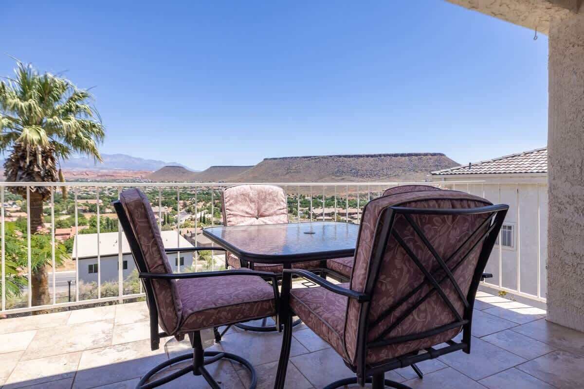A balcony with a table and chairs, overlooking a landscape with mountains and a palm tree.