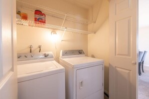 Une buanderie équipée de deux machines à laver et deux sèche-linge superposés, d’une étagère contenant des produits de nettoyage et d’une porte donnant sur une autre pièce.