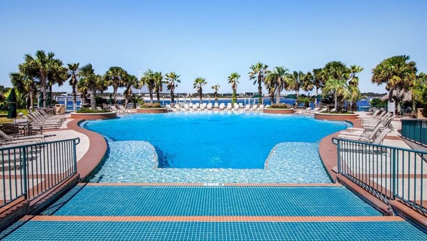 A large swimming pool with a clear blue water, surrounded by lounge chairs and palm trees. - Caribe Resort C712 | Lazy River, Pools, Hot Tubs, Gym, Tennis | Waterfront Condo | Beach Getaways (Orange Beach)