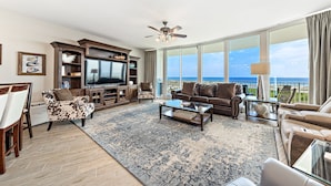 A spacious living room with a large flat-screen TV, comfortable seating, and a view of the ocean. - Caribe Resort C712 | Lazy River, Pools, Hot Tubs, Gym, Tennis | Waterfront Condo | Beach Getaways (Orange Beach)