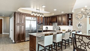 A modern kitchen with dark wood cabinets, a large island with bar stools, stainless steel appliances, and decorative wall clocks. - Caribe Resort C712 | Lazy River, Pools, Hot Tubs, Gym, Tennis | Waterfront Condo | Beach Getaways (Orange Beach)
