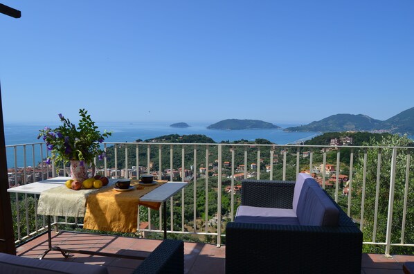 A balcony with a table set for dining, a wicker sofa, and a view of the sea and hills. - la castellana una finestra sul mare (Lerici)