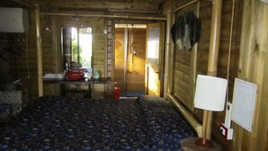 Une pièce de cabane en bois comprenant un lit, une petite table avec un bol rouge, une bouteille et un miroir. Une porte donne accès à une autre pièce.
