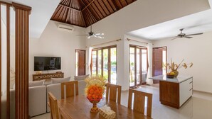 A spacious dining area with a wooden table, chairs, and a decorative centerpiece. There are ceiling fans, a television, and a kitchen area visible.
