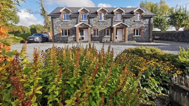 A stone building with multiple doors and windows, surrounded by greenery and a gravel driveway.