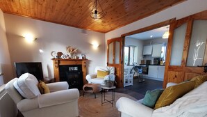 A cozy living room with two white sofas, a fireplace, a television, and a kitchen visible through an open doorway.