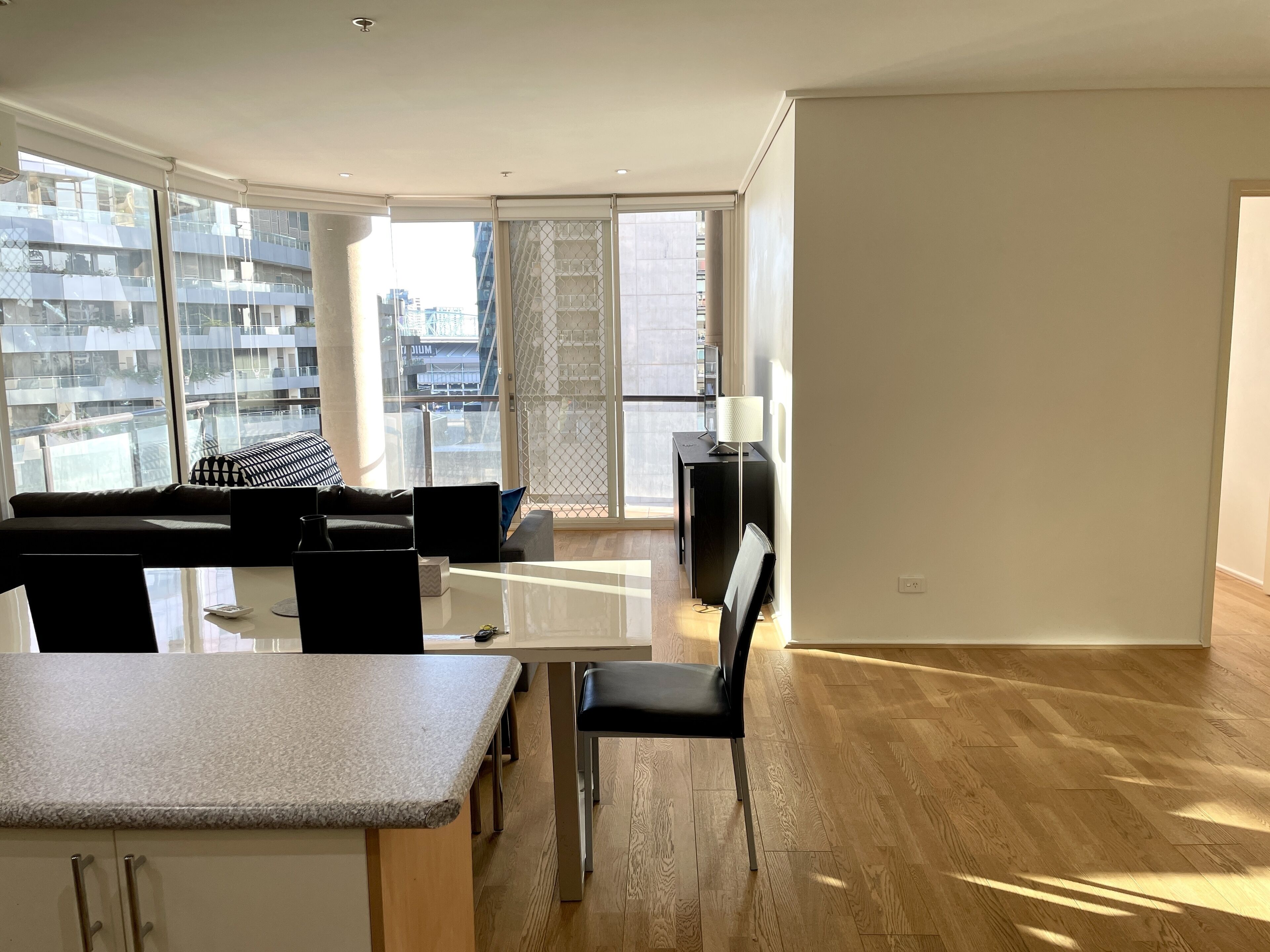 Un soggiorno moderno con un divano, un tavolo da pranzo e una TV. Ampie finestre offrono vista su edifici circostanti.