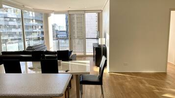 Un soggiorno moderno con un divano, un tavolo da pranzo e una TV. Ampie finestre offrono vista su edifici circostanti.