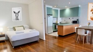 Quarto de hotel moderno com cama, uma pequena cozinha, área de jantar com mesa e cadeiras, e um quadro pendurado na parede.