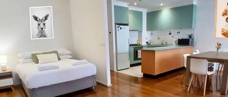 A modern hotel room with a bed, a small kitchenette, a dining area with a table and chairs, and a framed picture on the wall.