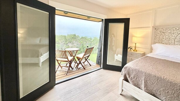 Un dormitorio con cama, mesita de noche, lámpara y vistas a un balcón con mesa y sillas.