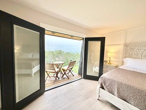 A bedroom with a bed, a nightstand, a lamp, and a view of a balcony with a table and chairs.