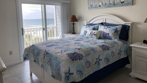 Quarto com cama, mesa de cabeceira, abajur, ventilador de teto, pintura de barcos à vela e vista para o oceano através de uma porta de correr.