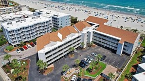 Vista aérea de uma área costeira com praia, estacionamento e vários edifícios.