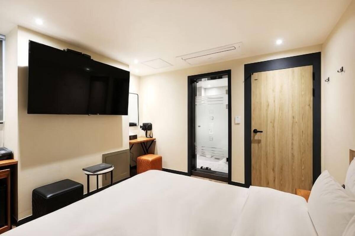 Ein Hotelzimmer mit einem Bett, einem Schreibtisch mit Computer, einem an der Wand montierten Fernseher und einer Holztür.