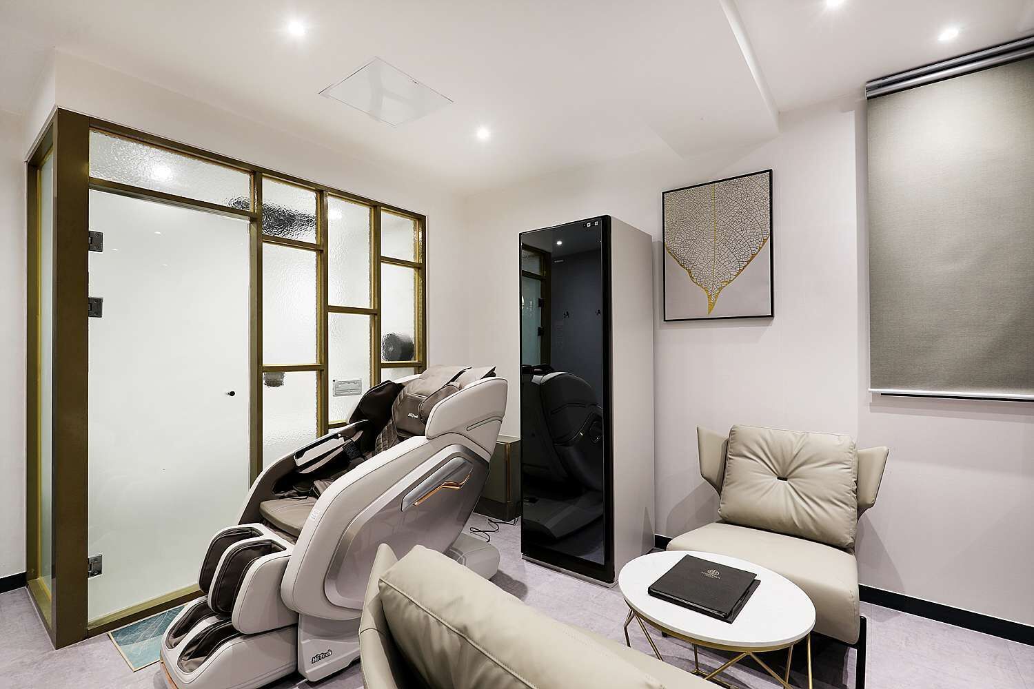 A modern room with a massage chair, a small round table with a book, a sofa, and a large abstract painting on the wall.