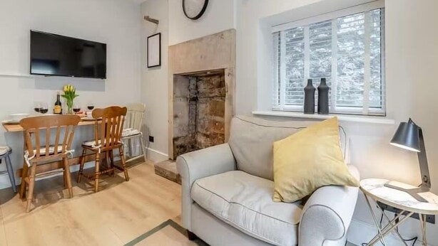 A living room with a sofa, a coffee table, a fireplace, a TV mounted on the wall, and a window with blinds. - Cottage in the heart of the Peak District (Matlock)