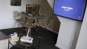Ein modernes Wohnzimmer mit einem runden Glastisch, Metallstühlen, einem Flachbildfernseher, der das Prime Video Logo zeigt, und einer Vase mit getrockneten Blumen.