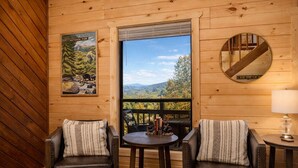 Une pièce chaleureuse aménagée comme une cabane, avec deux fauteuils en cuir, une petite table, une lampe et une grande fenêtre offrant une vue magnifique sur les montagnes et les arbres.