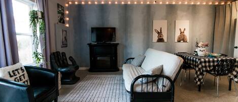 Una sala de estar con sofá, sillón y una mesa de comedor con sillas. Hay un televisor sobre un mueble bajo y una chimenea.