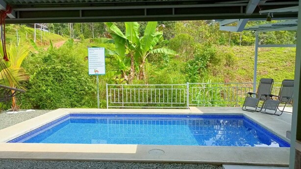 Una piscina rectangular al aire libre con superficie azul de azulejos, rodeada por una cerca metálica y un área techada con sillas.