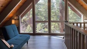 A room with a sloped ceiling, large windows, a blue sofa, and a wooden railing. - Tahoe Time Cabin w/King Bed, Modern Tahoe Cabin w/ King Bed Suite & Level 1 EV Charging (Incline Village)