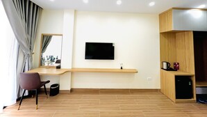 A modern living room with a wooden floor, a flat-screen TV mounted on the wall, a wooden coffee table, a dark chair, and a small trash bin. - Orion Hotel Halong (Hong Gai)