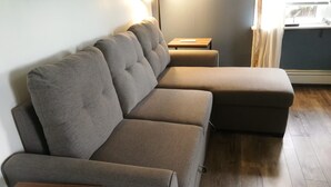 A sectional sofa with a wooden side table, a small table, and a window with curtains. - Cozy private cottage with picturesque scenery (New Glasgow)