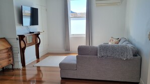 A modern living room with a grey sofa, wooden flooring, a flat-screen TV mounted on the wall, and a view of the outdoors through a window with curtains. - North Edge Cottage ~ Free Parking, City, Warm (Launceston)