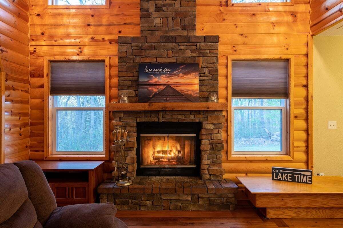L’intérieur d’un chalet en rondins, avec une cheminée en pierre, deux fenêtres équipées de stores, un coin salon confortable et un panneau indiquant « TEMPS POUR LE LAC ».