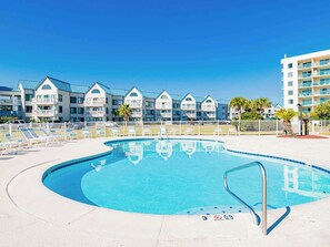 Una piscina con cubierta blanca, rodeada de una valla blanca, y una fila de casas de playa al fondo.