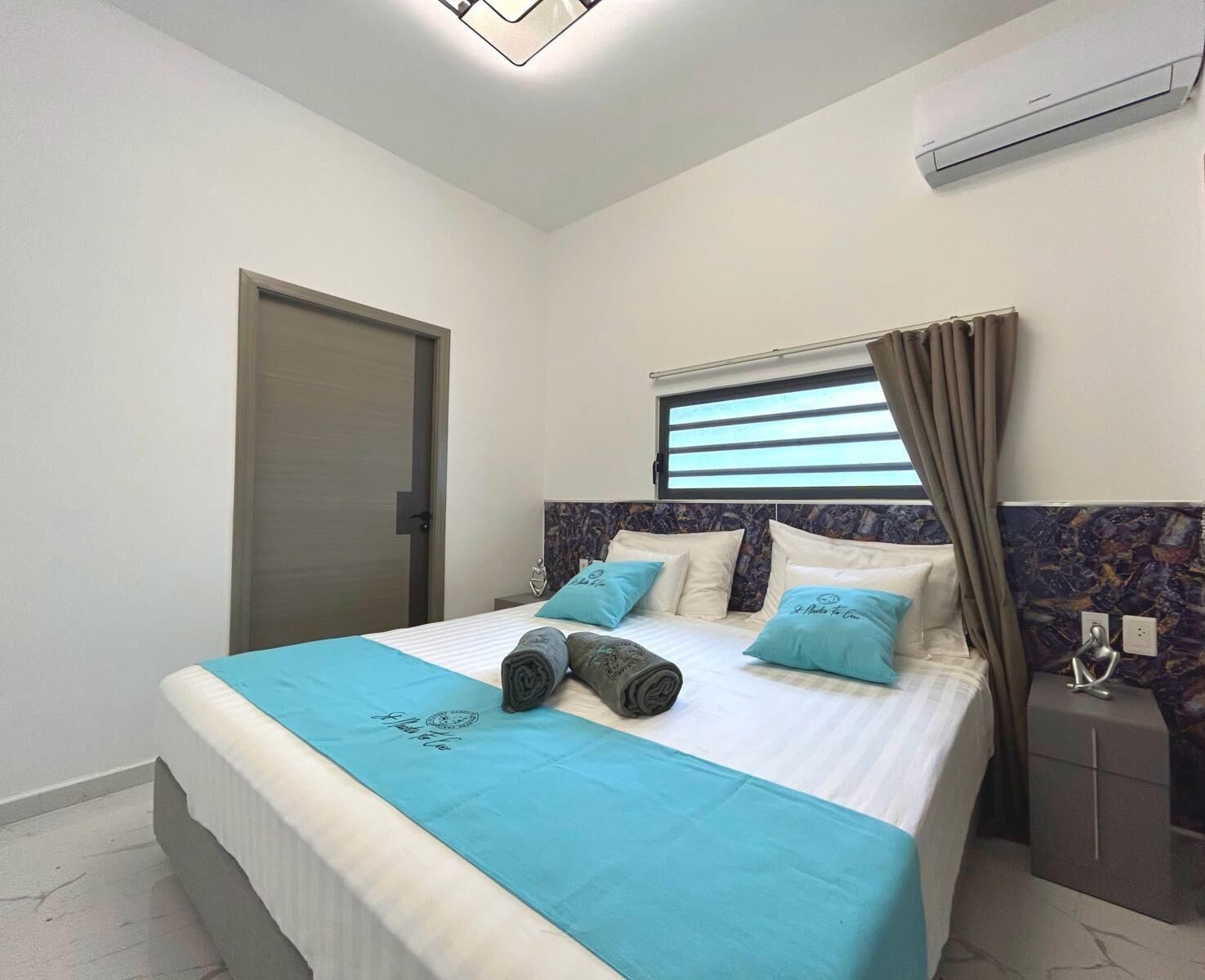 Un dormitorio con cama, mesita de noche, ventana con cortinas, aire acondicionado y puerta.