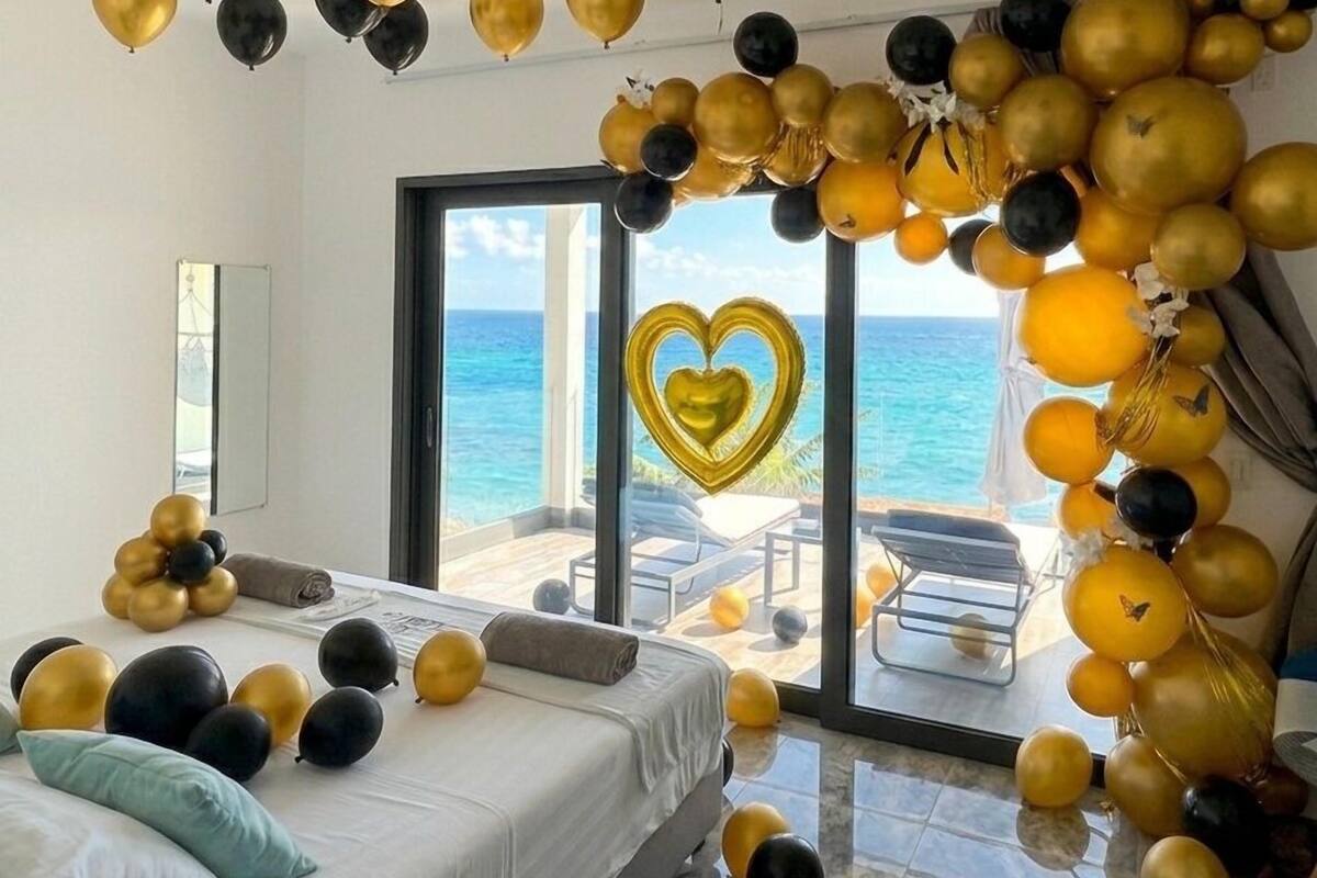 A room with a bed, a large window with a view of the sea, and balloons.