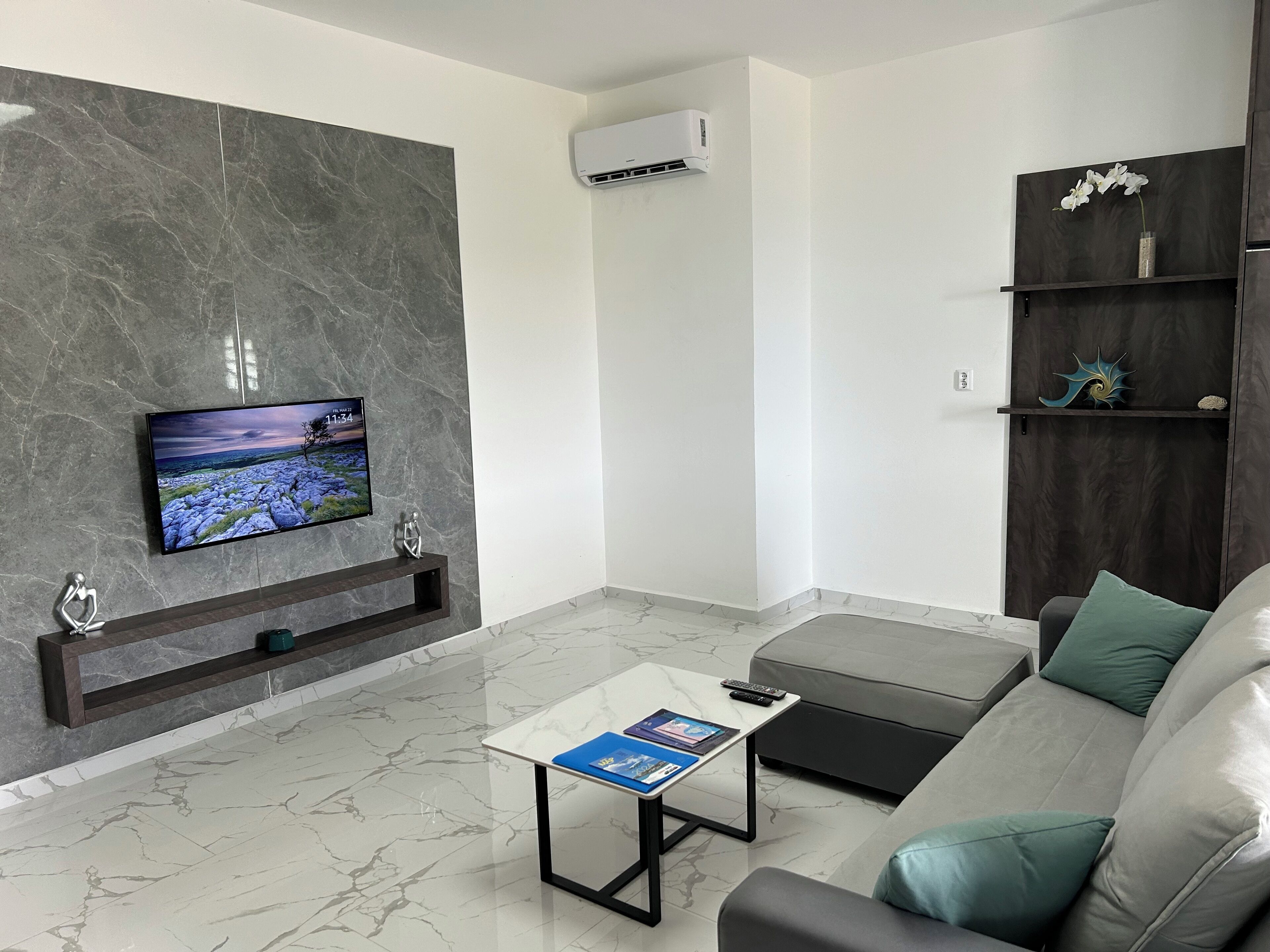 A modern living room with a marble floor, a grey sofa, a coffee table, a TV mounted on a marble wall, and a dark wooden shelf with decorative items.